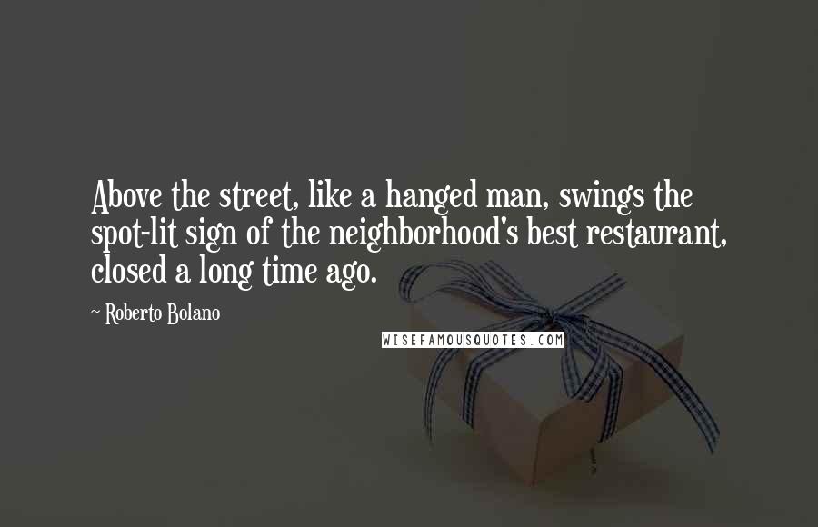 Roberto Bolano Quotes: Above the street, like a hanged man, swings the spot-lit sign of the neighborhood's best restaurant, closed a long time ago.